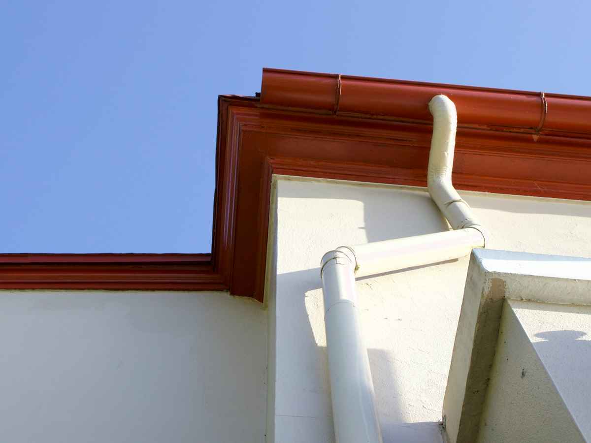 A brown gutter along a white roof edge running into a white downspout under a blue sky